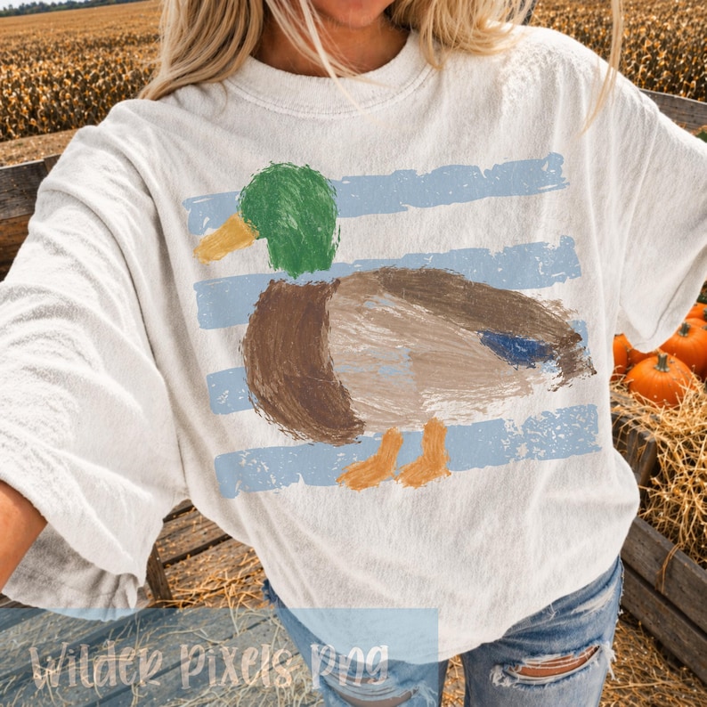 Pu&ograve; includere: T-shirt bianco sporco con un disegno di anatra dipinta, con testa verde, corpo marrone e becco e zampe gialle. L'anatra &egrave; posta su strisce orizzontali azzurre. Il testo "Wilder Pixels Png" &egrave; in basso.