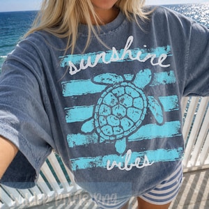 May include: A blue t-shirt with a turquoise turtle graphic and the words "sunshine vibes". The design includes horizontal stripes and the text is in a cursive font. The shirt is a relaxed fit.