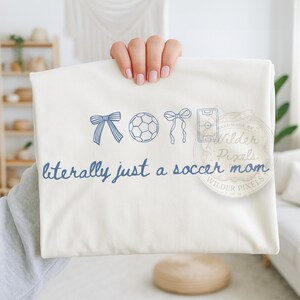 May include: Cream-colored t-shirt with blue soccer-themed graphics and text. The design includes a bow, soccer ball, another bow, and a soccer field. The text reads "literally just a soccer mom."
