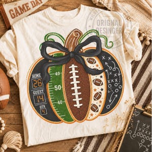 May include: A cream-colored t-shirt featuring a graphic of a pumpkin designed like a football. The pumpkin includes a scoreboard reading Home 28, Guest 14, Qtr 4, a green field section with yard lines, a brown football center, and a black bow.