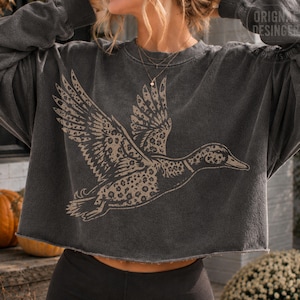 Puede incluir: Una sudadera corta de color gris carb&oacute;n con un gr&aacute;fico grande de estilo desgastado de un pato volando con un patr&oacute;n de leopardo beige y marr&oacute;n. La prenda tiene mangas largas, cuello redondo y un dobladillo sin rematar en la cintura.