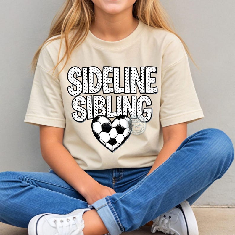 May include: A beige t-shirt with the words "SIDELINE SIBLING" in a black dotted font. A heart-shaped soccer ball graphic is centered below the text. The person is wearing blue jeans and white sneakers.