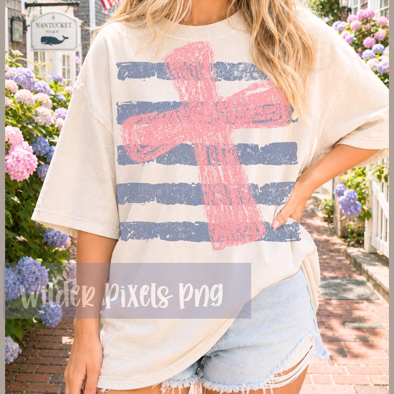 May include: Cream-colored t-shirt featuring a pink cross design over blue horizontal stripes. The shirt has a relaxed fit and short sleeves. The text "Wilder Pixels Png" is visible at the bottom of the shirt.