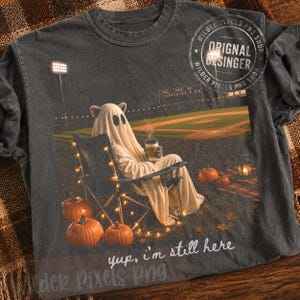 May include: A charcoal gray t-shirt featuring a graphic of a ghost wearing cat ears, sitting in a folding chair on a baseball field at night, holding a coffee cup. The scene includes pumpkins, string lights, and the text, "yup, i'm still here."