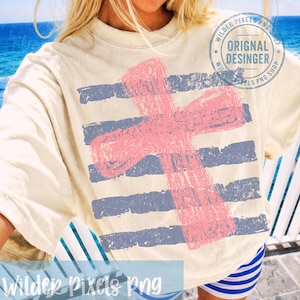 May include: Cream-coloured t-shirt with a distressed pink cross design over blue horizontal stripes. The shirt has a vintage, worn look. The text "Wilder Pixels Png" is at the bottom of the shirt. The shirt also has a circular stamp that says "Original Desinger".