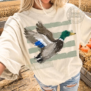 May include: A cream-colored t-shirt featuring a painterly graphic of a mallard duck in flight. The duck has a green head, yellow beak, and blue wing patch. Behind the duck are three horizontal, distressed sage green brushstroke stripes.