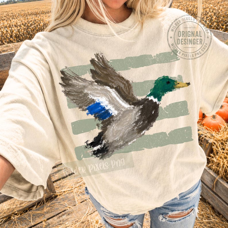 May include: A cream-colored t-shirt featuring a painterly graphic of a mallard duck in flight. The duck has a green head, yellow beak, and blue wing patch. Behind the duck are three horizontal, distressed sage green brushstroke stripes.