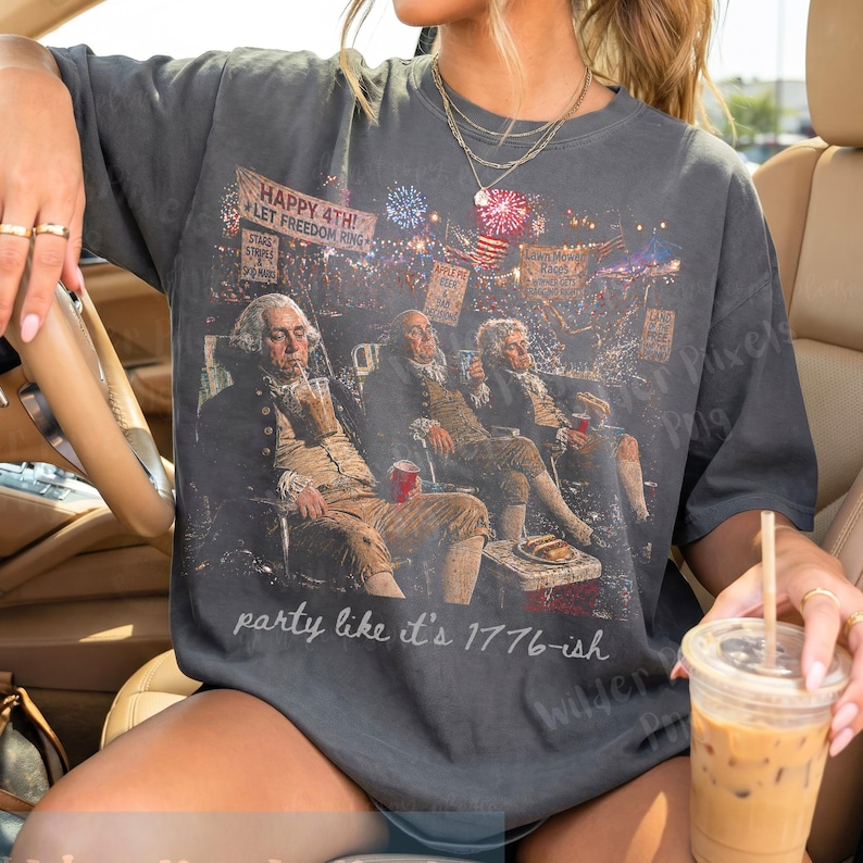 May include: Dark gray t-shirt featuring a graphic of historical figures enjoying a fireworks display. The shirt has the text "HAPPY 4TH! LET FREEDOM RING" and "party like it's 1776-ish". The design includes red, white, and blue colors.