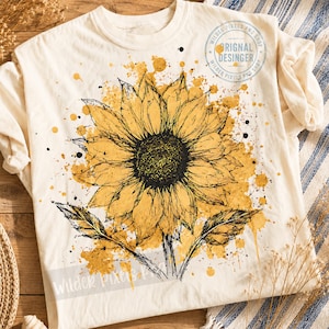 May include: A cream-colored t-shirt featuring a large, detailed black ink sketch of a sunflower centered on the chest. The flower is layered over a vibrant yellow watercolor-style paint splatter design. Text in the upper right reads ORIGINAL DESINGER.