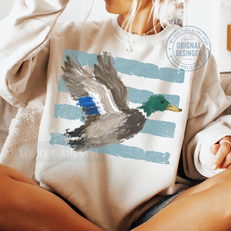 May include: A beige crewneck sweatshirt featuring a painterly graphic of a mallard duck in flight. The duck has a green head, grey body, and blue wing patch. Behind the duck are horizontal light blue brushstroke stripes. A circular watermark is visible.