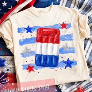 May include: A cream-coloured t-shirt with a red, white, and blue popsicle design. The shirt features blue and white painted stripes and scattered stars in red, blue, and silver. A patriotic theme.