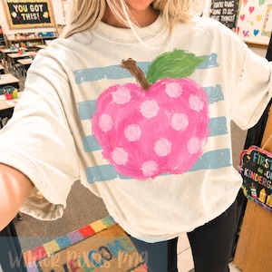 Puede incluir: Camiseta color crema con una gran manzana rosa con lunares blancos, un tallo marrón y una hoja verde. Rayas horizontales de color azul claro están pintadas detrás de la manzana. El texto "YOU GOT THIS!" es visible en el fondo.