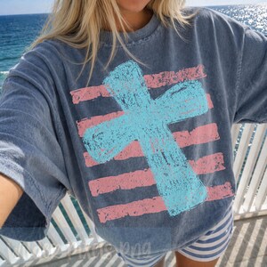 May include: A blue t-shirt with a large, light blue cross design over pink horizontal stripes. The shirt has a slightly faded, vintage look. The overall theme is religious or spiritual.