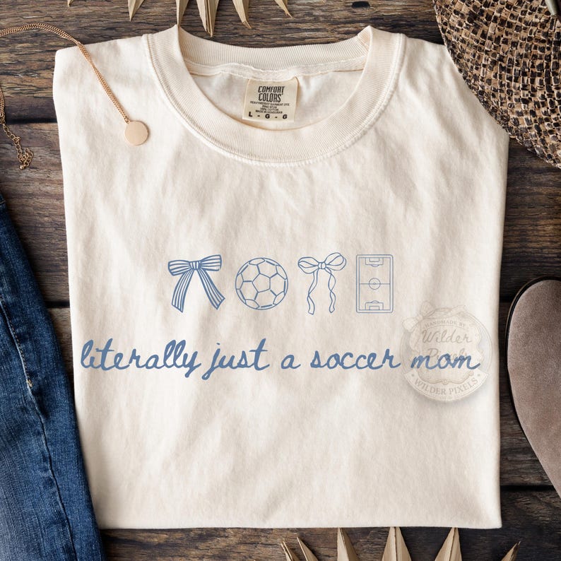 May include: Cream-colored t-shirt with blue graphic elements and text. The design includes a bow, soccer ball, another bow, and a soccer field. The text reads "literally just a soccer mom."