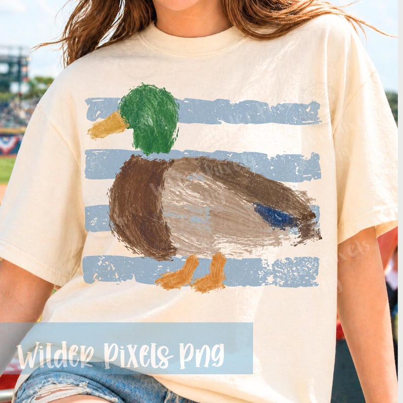 Pu&ograve; includere: T-shirt color crema con un disegno di anatra dipinta e strisce blu. L'anatra ha la testa verde, il corpo marrone e le zampe arancioni. Il testo "Wilder Pixels Png" &egrave; in basso.