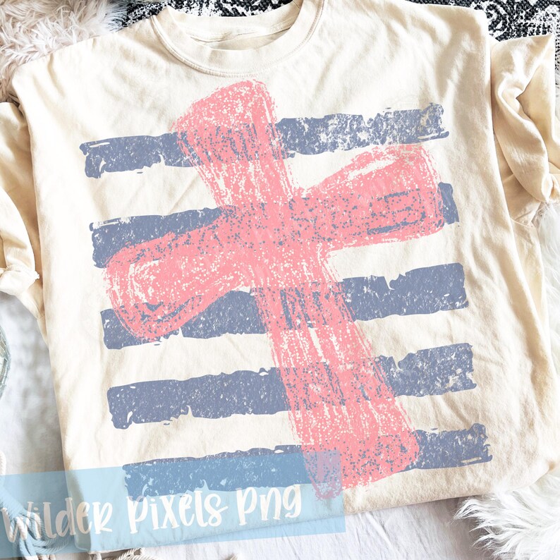 May include: Cream-colored t-shirt featuring a distressed pink cross design over blue horizontal stripes. The graphic has a textured, worn appearance. The shirt is made of soft material.