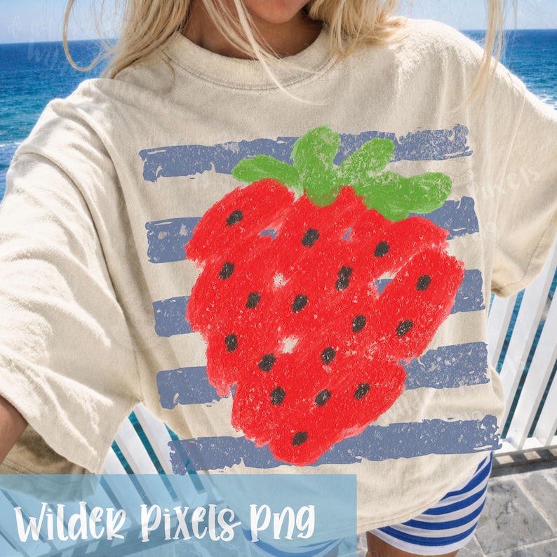 Op de afbeelding: Cr&egrave;mekleurig T-shirt met een grote, rode aardbeienillustratie met groene bladeren en zwarte zaden. Het ontwerp bevat blauwe horizontale strepen. De tekst "Wilder Pixels Png" staat onderaan.