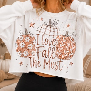 May include: A white long-sleeve cropped sweatshirt featuring a graphic print of three pumpkins in orange and brown tones. The text "I love Fall The Most" is centered in brown script. The pumpkins are decorated with floral and leaf patterns.