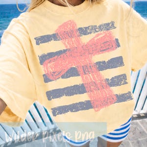 May include: Yellow short-sleeved shirt with a distressed pink cross design over horizontal gray stripes. The shirt is worn with blue and white striped shorts. The background shows a glimpse of the ocean.