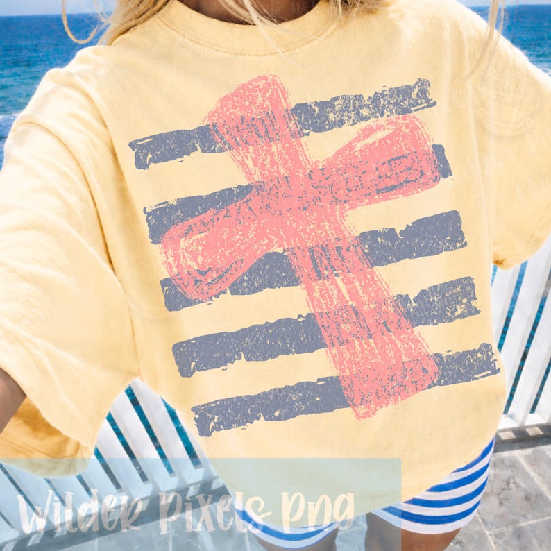 May include: Yellow short-sleeved shirt with a distressed pink cross design over horizontal gray stripes. The shirt is worn with blue and white striped shorts. The background shows a glimpse of the ocean.
