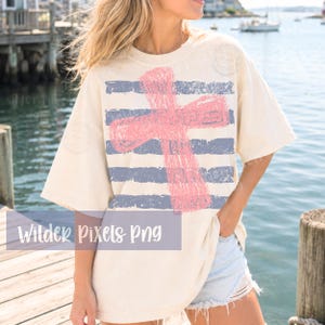 May include: Cream-colored t-shirt featuring a distressed pink cross over blue and white horizontal stripes. The shirt has a relaxed fit and short sleeves. The text "Wilder Pixels Png" is visible at the bottom of the image.