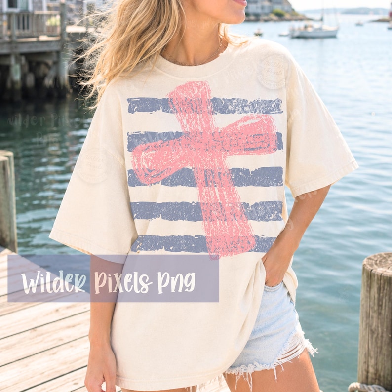 May include: Cream-colored t-shirt featuring a distressed pink cross over blue and white horizontal stripes. The shirt has a relaxed fit and short sleeves. The text "Wilder Pixels Png" is visible at the bottom of the image.