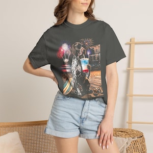 May include: A charcoal gray short-sleeve t-shirt featuring a graphic print of a spotted dog sitting on a wooden deck at night. The dog holds a red, white, and blue layered drink, with colorful fireworks exploding in the dark sky above the beach.