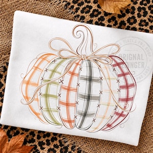 May include: A white t-shirt featuring a large graphic of a pumpkin composed of vertical segments in orange, green, and red plaid patterns. The pumpkin is outlined with brown line art and includes a decorative bow at the top stem with cross-stitch details.