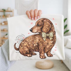 Può includere: Una maglietta color crema con un'illustrazione di un cane bassotto marrone, dettagli glitterati dorati e un papillon color crema con pois neri. Cuori rosa circondano il cane. Il testo "Wild Pixels Pizza" è in un design circolare.