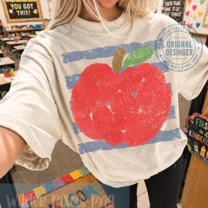Puede incluir: Camiseta color crema con un gran gráfico de manzana roja desgastada con una hoja verde y un tallo marrón. La manzana está sobre rayas horizontales azules. El texto "YOU GOT THIS!" es visible en el fondo.
