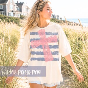 May include: Cream-colored t-shirt featuring a large pink cross design over blue horizontal stripes. The shirt has a relaxed fit and short sleeves. The text "Wilder Pixels Png" is visible at the bottom of the image.