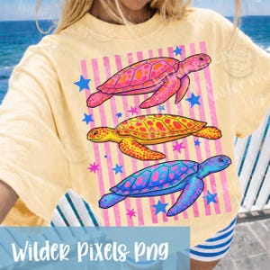 May include: Yellow t-shirt featuring a graphic of three colorful sea turtles. The turtles are pink, yellow, and blue, set against a pink striped background with blue and pink stars. The text "Wilder Pixels Png" is at the bottom.