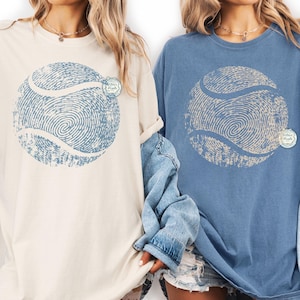 May include: Two long-sleeve t-shirts, one cream and one blue, each featuring a large tennis ball design made of fingerprint patterns. The shirts have a relaxed fit and are suitable for casual wear. The text "Wilder Pixels" is on the shirts.