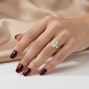 May include: A gold ring with a rectangular, emerald-cut diamond center stone, surrounded by smaller diamonds. The ring is on a hand with burgundy nail polish, set against a neutral background.