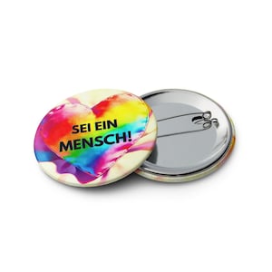 Könnte beinhalten: Zwei runde Ansteckbuttons. Ein Button zeigt ein buntes Aquarellherz mit dem schwarzen Text SEI EIN MENSCH! Der andere Button liegt mit der silbernen Metallrückseite nach oben und zeigt den Verschlussmechanismus zum Befestigen an Kleidung.