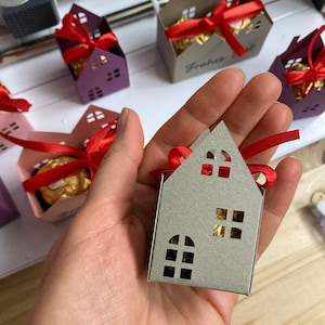 May include: A small, gray house-shaped gift box with cut-out windows and a red ribbon. Other boxes in purple and brown are visible, each with a red ribbon and a gold-wrapped treat inside. The boxes are on a white surface.