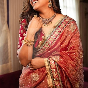 May include: A woman wearing a red sari with gold embroidery and embellishments. The sari has a textured appearance and is paired with gold jewellery, including a necklace, earrings, and bangles. The sari's border is detailed with gold trim.