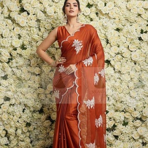 May include: Rust-colored saree with white floral embroidery and a scalloped edge. The sleeveless blouse has a V-neckline. The saree features a sheer, embroidered border and is draped over the body. The background is a wall of white roses.