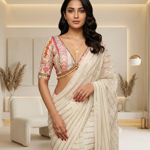 Puede incluir: Una mujer con un sari blanco con rayas doradas y una blusa bordada colorida con un escote en V profundo. Lleva joyas de oro, incluyendo un collar y anillos. El fondo es una habitación de tonos neutros con una silla y una lámpara.