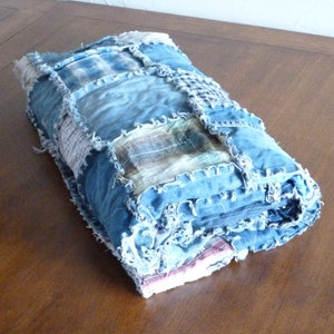 Op de afbeelding: Een blauwe denim quilt gemaakt van hergebruikt denim en geruit stof. De quilt is opgerold en heeft een rafelige rand.