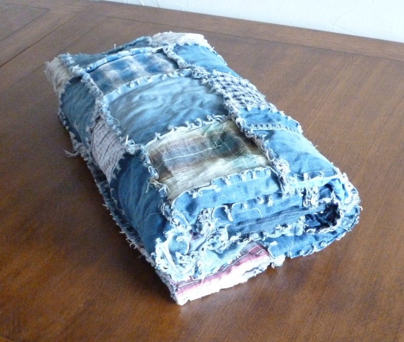Op de afbeelding: Een blauwe denim quilt gemaakt van hergebruikt denim en geruit stof. De quilt is opgerold en heeft een rafelige rand.