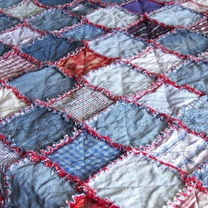 Op de afbeelding: Een patchworkdeken gemaakt van denim en andere stoffen in tinten blauw, rood en wit. De deken is in een ruitpatroon gestikt en heeft een rafelige rand.