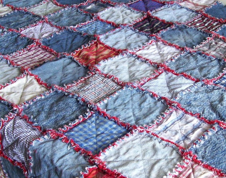 Op de afbeelding: Een patchworkdeken gemaakt van denim en andere stoffen in tinten blauw, rood en wit. De deken is in een ruitpatroon gestikt en heeft een rafelige rand.
