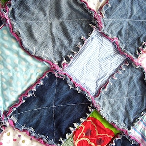 Op de afbeelding: Een patchworkdeken gemaakt van denimstof met verschillende gekleurde stofvierkanten. De deken is met roze draad aan elkaar genaaid en heeft een rafelige rand.