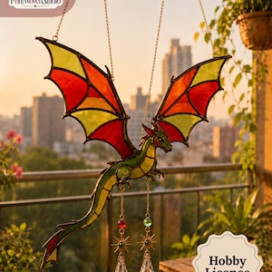 May include: A stained glass dragon suncatcher featuring green, red, and yellow glass panels held by dark metal cames. The dragon is suspended by two gold chains and features two dangling crystal prisms with gold star-shaped accents at the base.