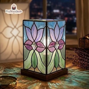 May include: A rectangular stained glass table lamp featuring a floral pattern with purple petals, green leaves, and light blue accents. The lamp has a dark wooden base and is illuminated from within, casting colorful light patterns on the surface.