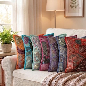 Puede incluir: Una colección de cojines decorativos en varios colores, incluyendo naranja, turquesa, rosa y morado. Los cojines presentan intrincados diseños de patchwork con detalles bordados y adornos. Están colocados sobre un sofá crema, creando un ambiente acogedor.