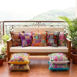 Puede incluir: Un sofá de exterior de madera con cojines color crema está adornado con una variedad de cojines decorativos de patchwork coloridos. Se apilan cojines adicionales en el suelo. Los cojines presentan patrones vibrantes en tonos naranja, morado, rosa y verde.