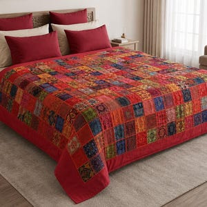 May include: A bedspread featuring a patchwork design of multicolored square fabric panels in shades of red, blue, yellow, and green. The quilt is draped over a bed with two solid red rectangular pillows and two solid cream rectangular pillows.