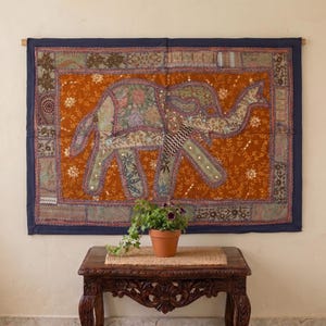May include: A rectangular wall hanging featuring a central orange panel with a large elephant appliqué. The elephant is decorated with various floral patterns and small mirrors. The piece is framed by a dark blue border with patchwork details.